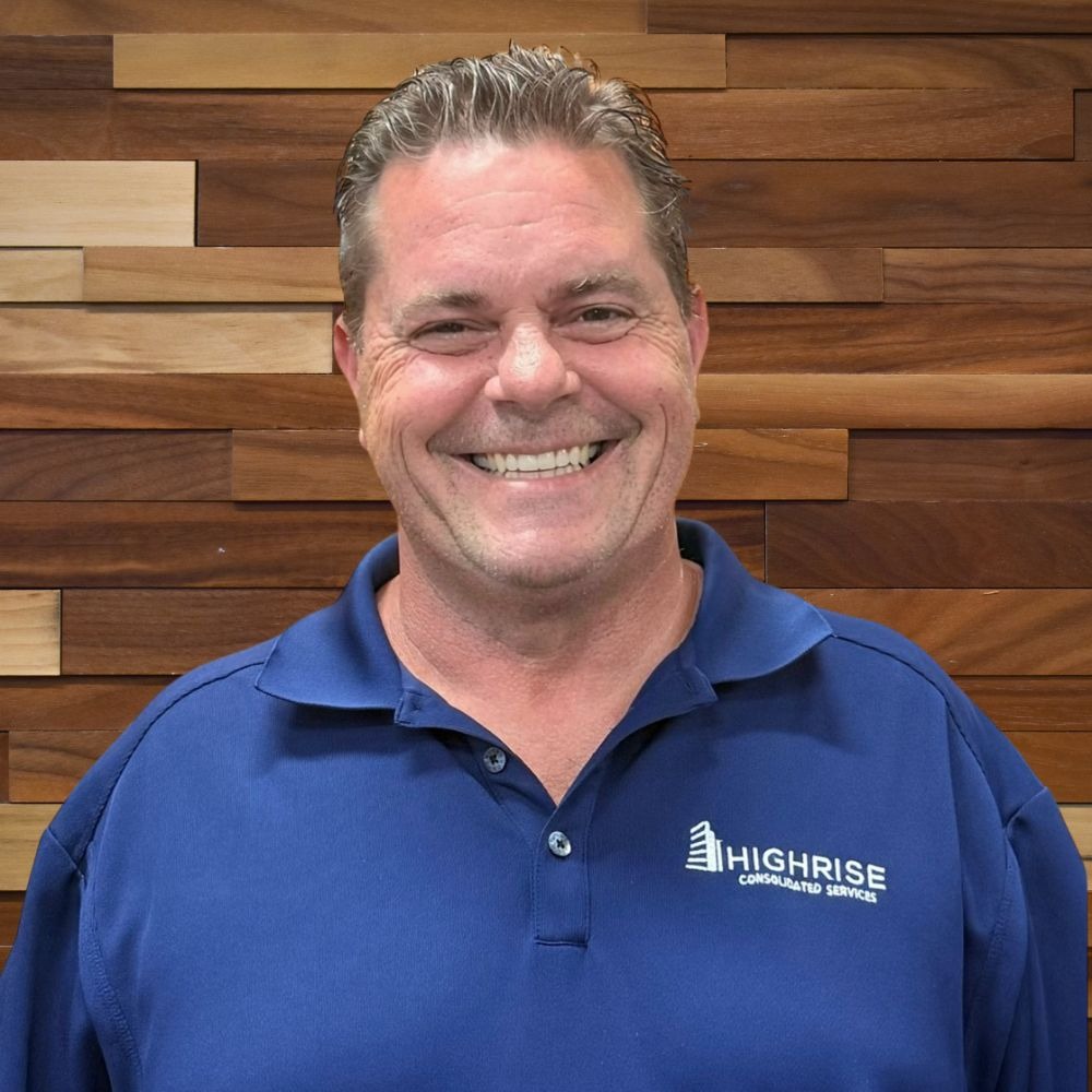 Smiling man in a blue polo shirt with Highrise Consolidated Services logo, standing in front of a wooden panel wall.