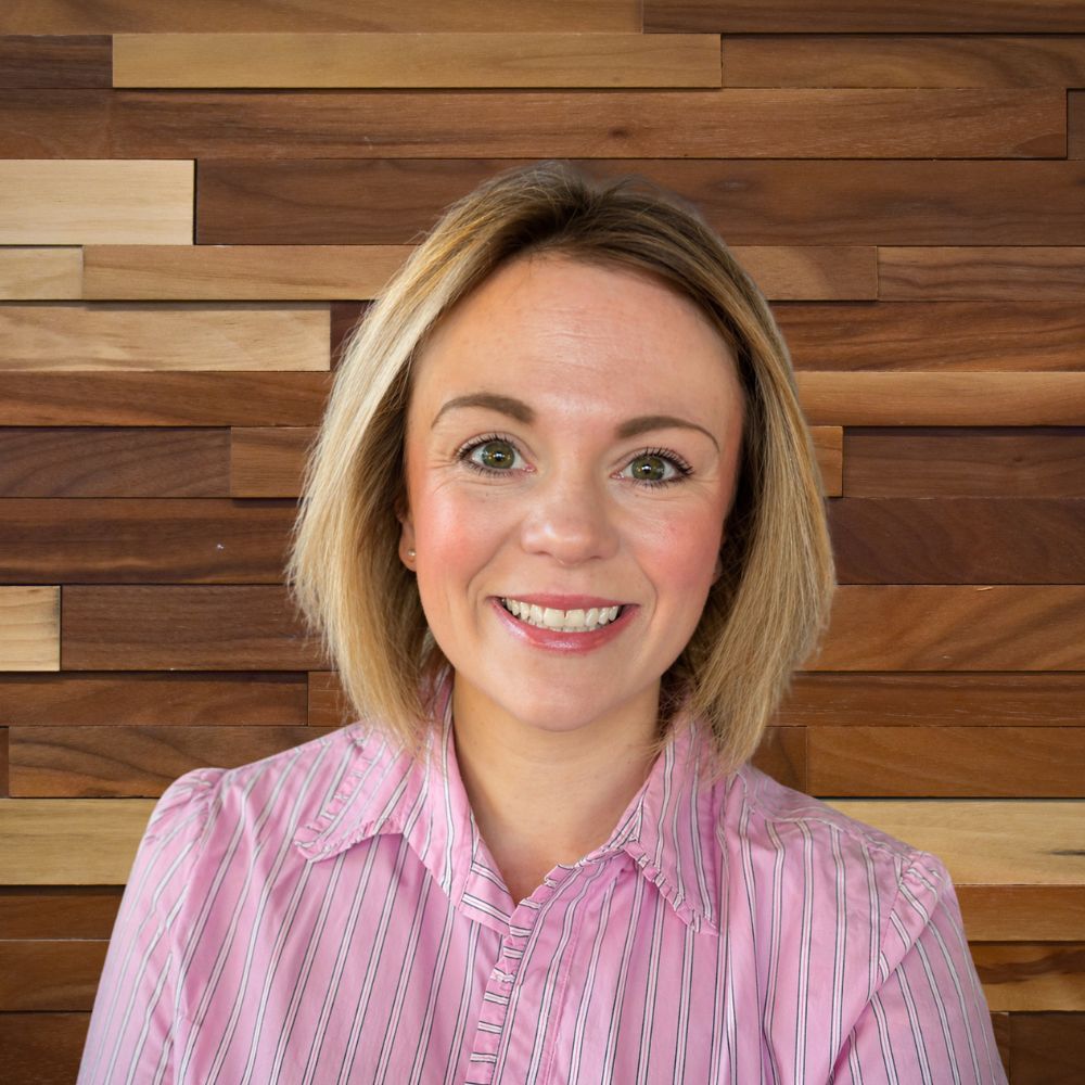 Smiling woman with short blonde hair in a pink striped blouse posing against a wooden panel wall.